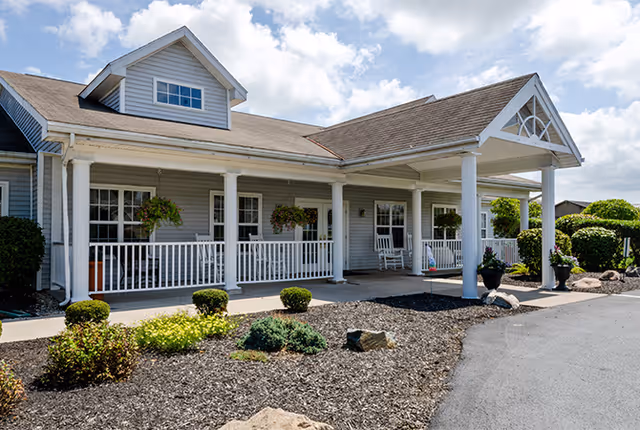 Covered front entrance and porch of a single-story senior living facility with white columns, hanging plants, shrubs, and a paved driveway.