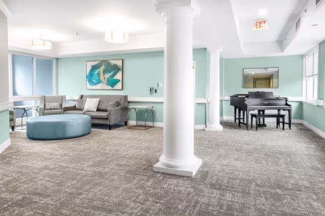 A spacious, well-lit common area with light green walls and large windows. The room features a gray sofa with pillows, a round blue ottoman, two small side tables, and a black grand piano with a matching bench. White columns and a beige carpet complete the room's decor.