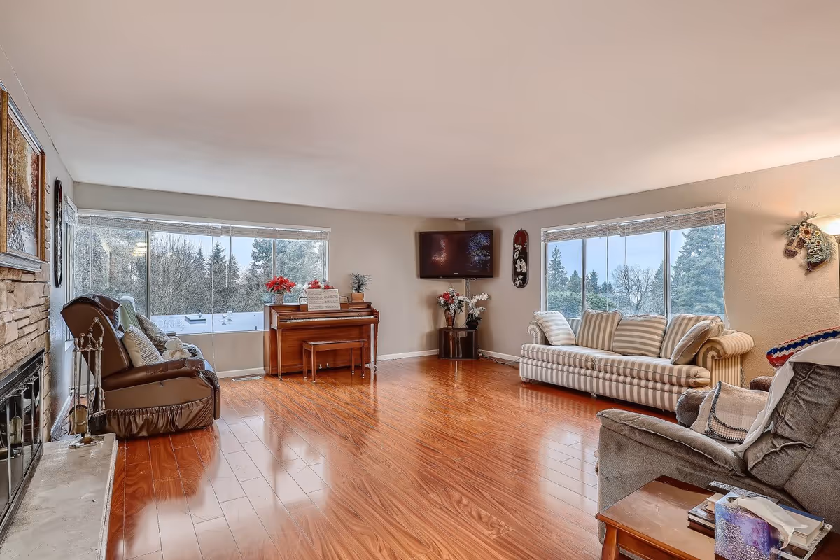 A spacious living room with polished wooden floors, a stone fireplace on the left, a brown leather armchair, a striped sofa, a gray recliner, a wooden piano with a bench, and large windows showing trees outside. A flat-screen TV is mounted on the wall above a small table with flowers.