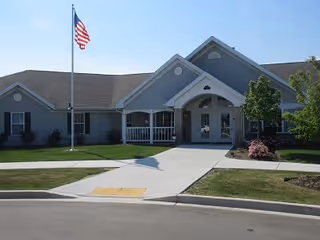 Exterior view of a single-story building with gray siding and a gabled roof, featuring a covered entrance with double doors. An American flag is flying on a flagpole in front of the building, and there are small trees and shrubs planted around the entrance area.