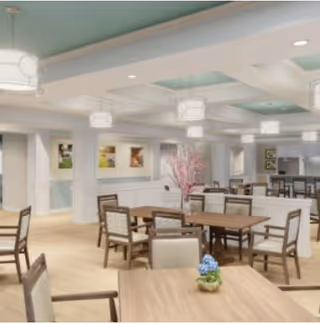 A spacious dining room with multiple wooden tables and chairs arranged neatly. The room features light-colored walls with white wainscoting, decorative ceiling lights, and pastel blue ceiling accents. There are framed pictures on the walls and a floral centerpiece on one of the tables.