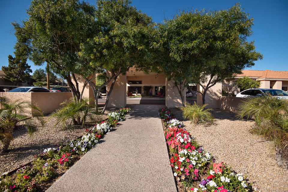 Pathway lined with colorful flowers leading to the entrance of a building with a covered porch, surrounded by trees and desert landscaping with gravel and small palm plants under a clear blue sky.