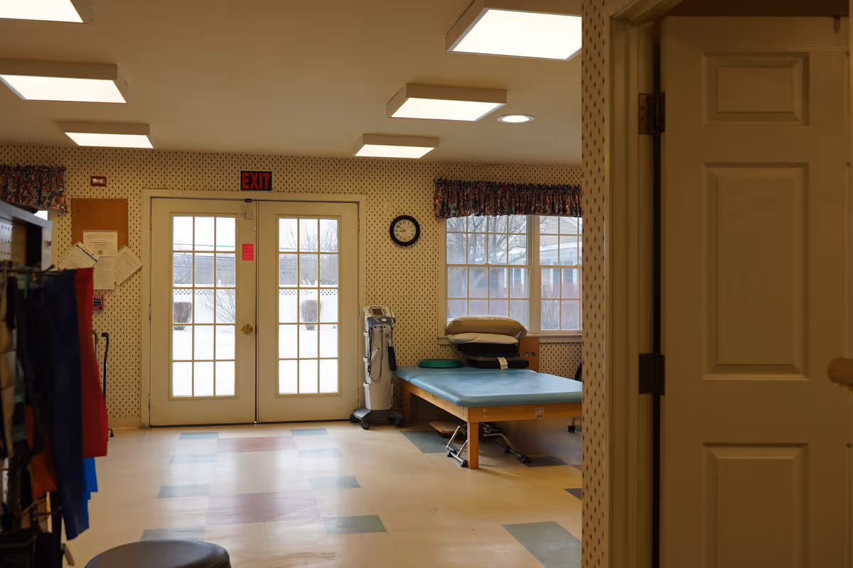 Interior room with patterned wallpaper, a treatment table with pillows, medical equipment on a stand, a clock on the wall, and double glass doors leading outside. The floor has a tiled pattern and there are fluorescent ceiling lights.