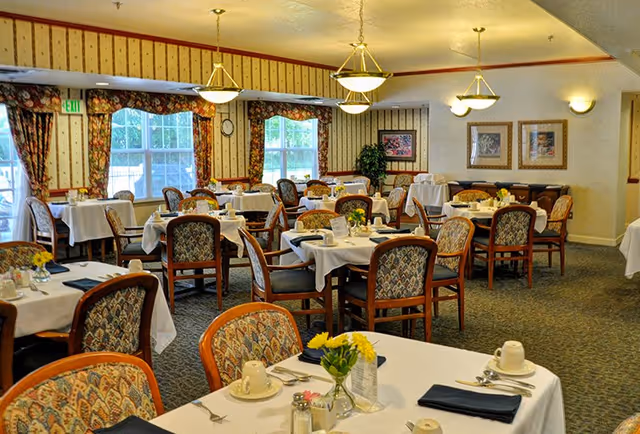 A bright dining room with multiple tables covered in white tablecloths, set with place settings and small floral centerpieces.