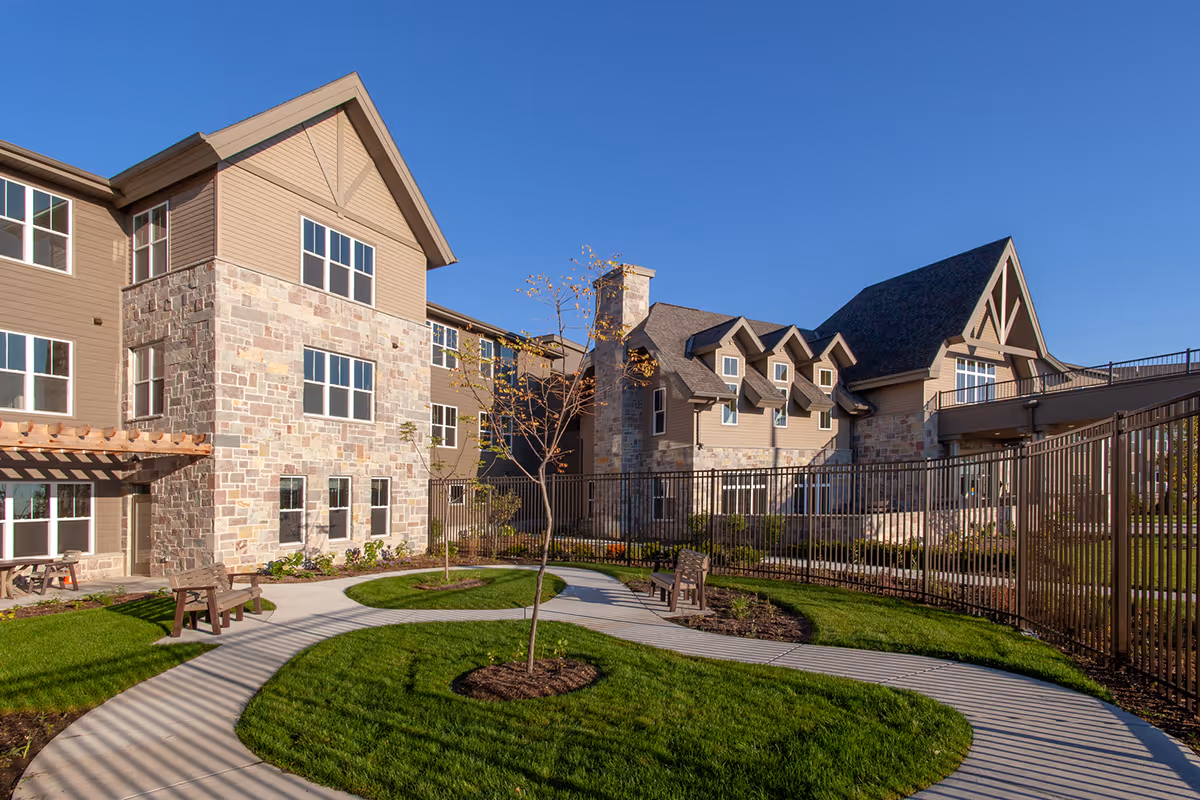 Outdoor courtyard area of a senior living facility with a winding concrete pathway, green grass, small trees, benches, and a multi-story building with stone and beige siding under a clear blue sky.