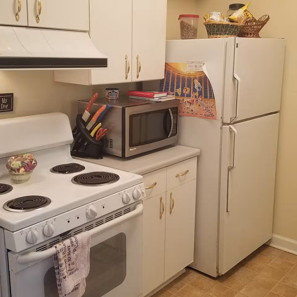 A small kitchen area with a white electric stove, a microwave on the countertop, white cabinets with gold handles, and a white refrigerator. There is a knife block with colorful knives next to the microwave, a small decorative bowl on the stove, and a poster on the refrigerator door. A basket with snacks and a container are on top of the refrigerator.