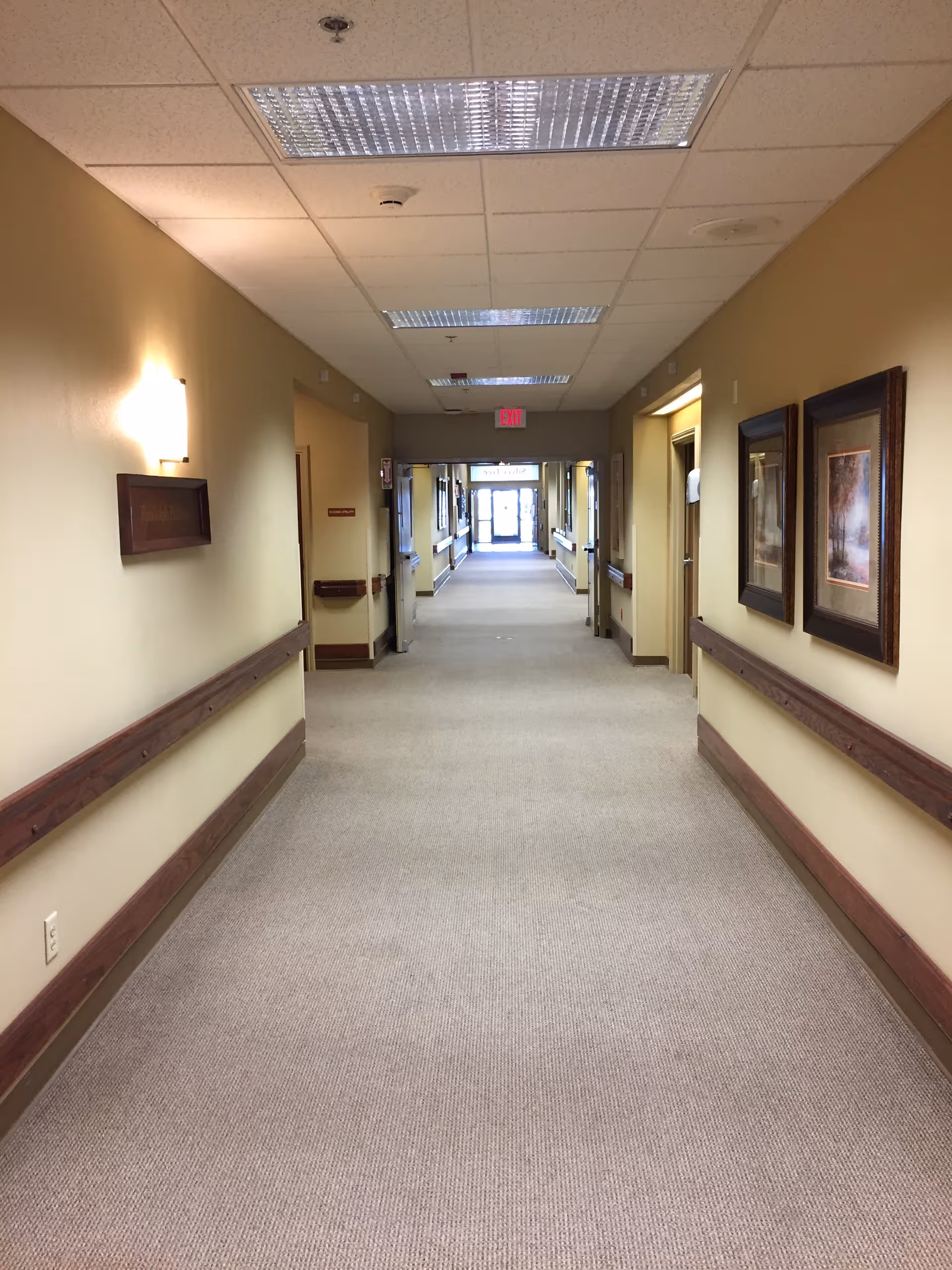 A long, well-lit hallway in a nursing and rehabilitation facility with beige walls, carpeted floor, wooden handrails on both sides, framed pictures on the right wall, and ceiling lights. The hallway leads to a glass exit door at the far end.