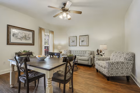A cozy living and dining area with a wooden dining table and four chairs in the foreground. The room features a patterned armchair, a light gray tufted sofa, two table lamps, framed bird artwork on the walls, a ceiling fan with lights, and hardwood flooring.