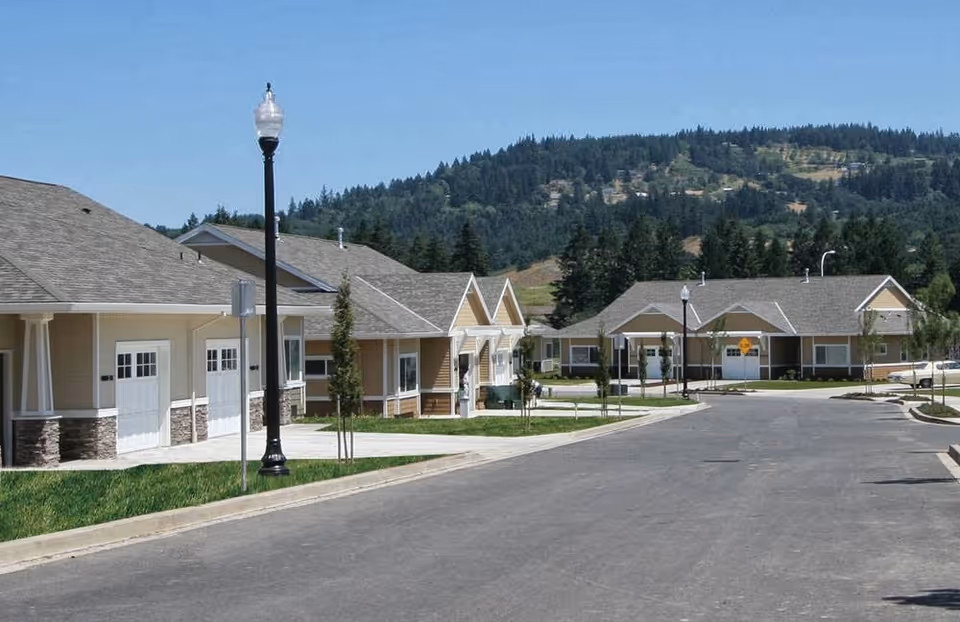 Single-story retirement cottages with garages and lampposts lining a paved street and forested hills in the background.