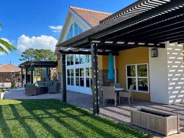 Outdoor patio area of a retirement community with pergolas casting shadows on the grass and paved floor. There are tables and chairs arranged for seating, with umbrellas folded and attached to some tables. The building has large windows and a sloped roof with tiles, under a clear blue sky with some clouds.