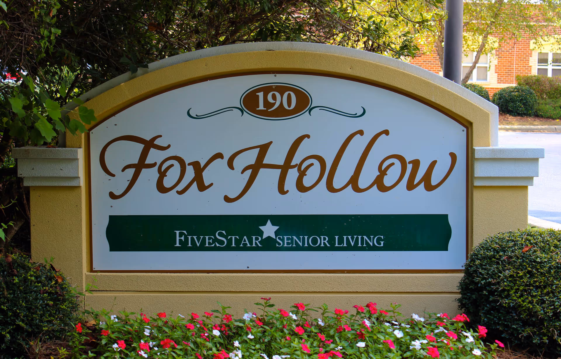 A large outdoor sign for Fox Hollow Senior Living Community, featuring the address number 190 and the Five Star Senior Living logo, surrounded by greenery and flowers.
