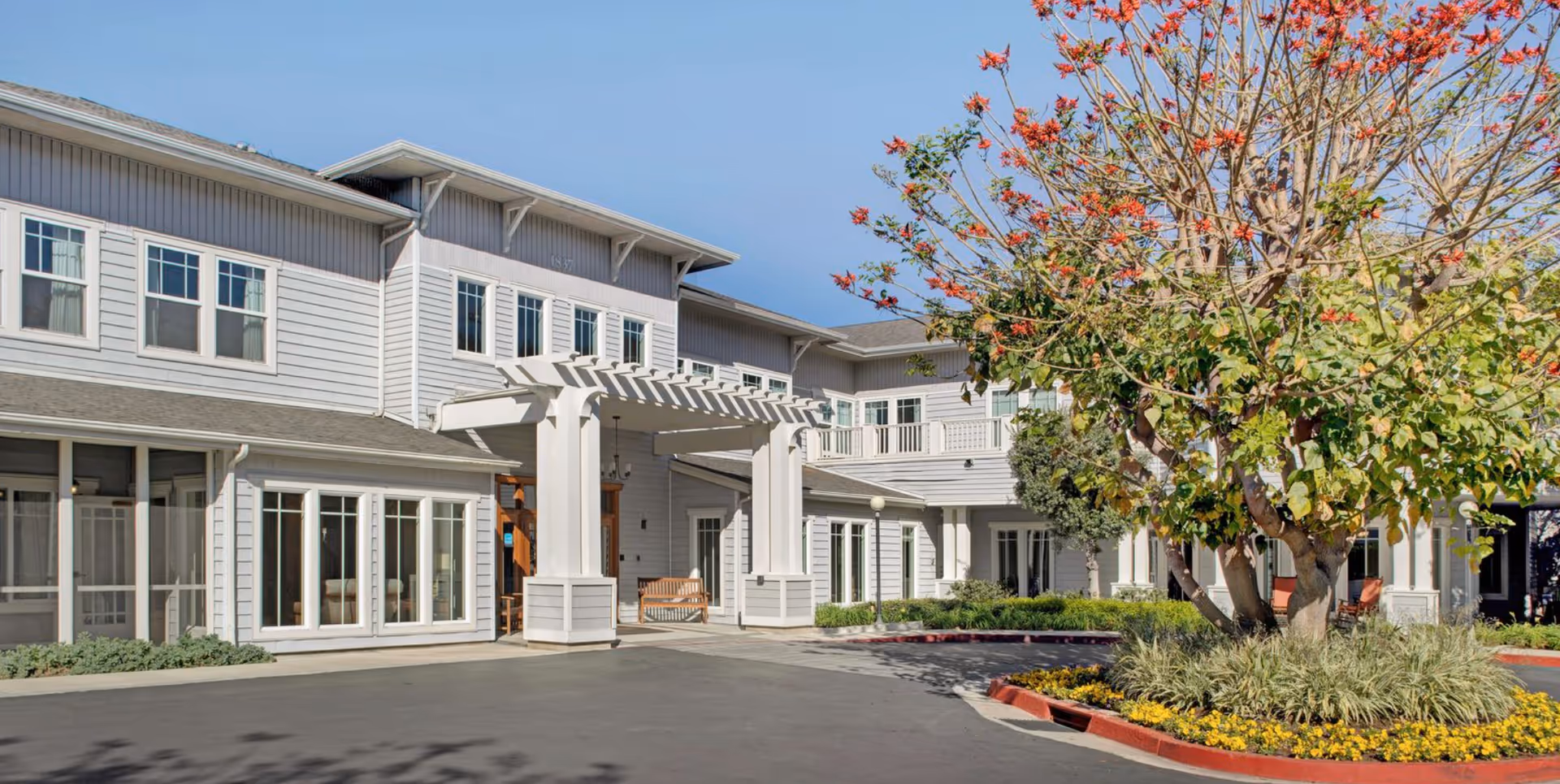 Front entrance of a two-story light-gray senior living building with a covered porte-cochere, a circular landscaped planter and a tree.