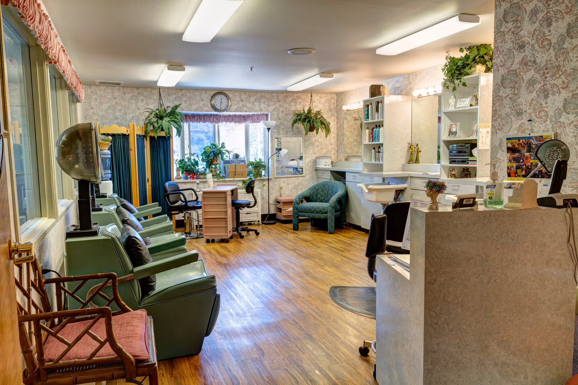 Salon-style common room with multiple green styling chairs and hooded hair dryers, a reception counter, sinks, and potted plants.