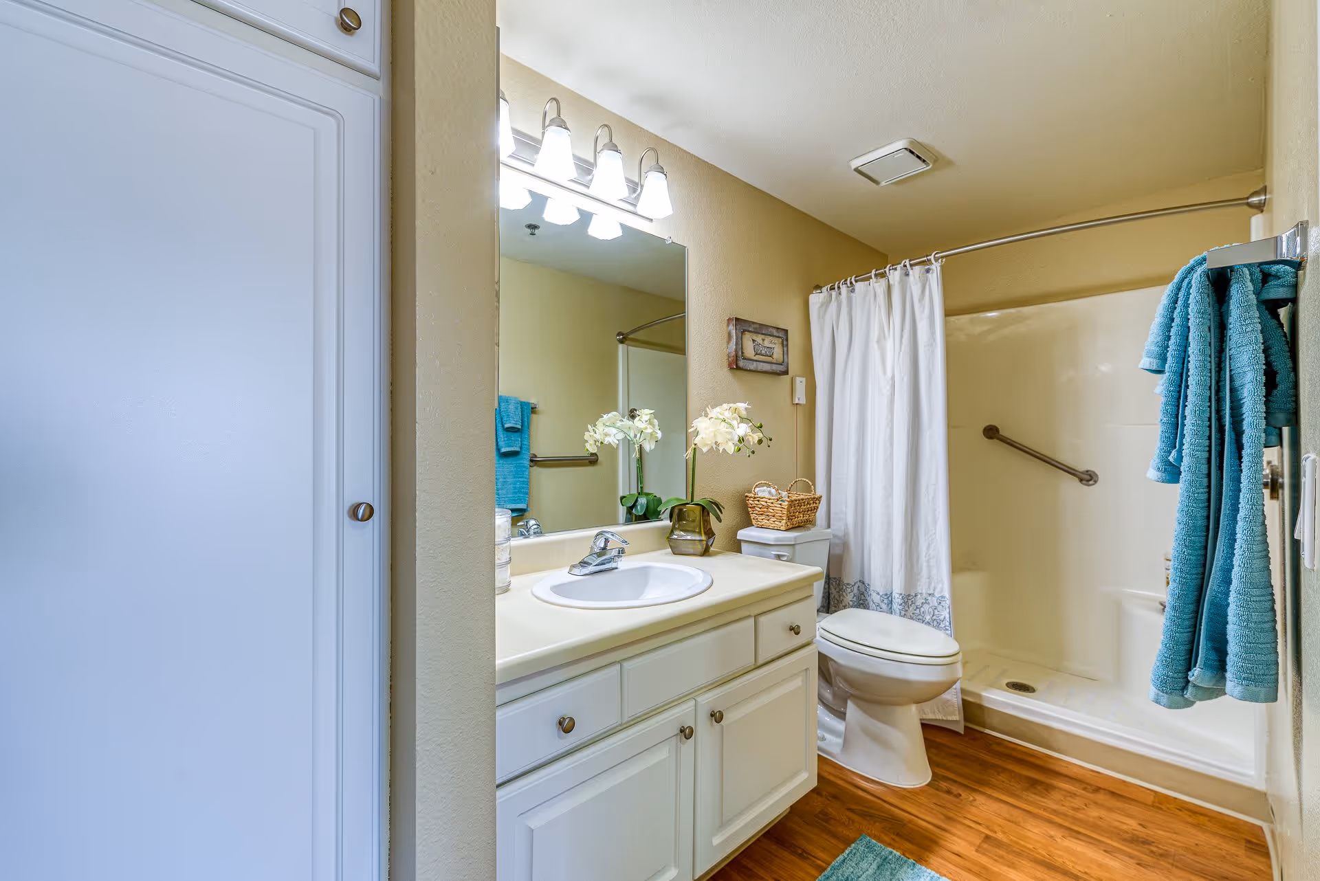 A clean and well-lit bathroom with a white vanity and sink, a large mirror with lights above it, a toilet, and a shower with a white curtain. There are teal towels hanging on a rack and a small basket on the toilet tank. The floor has a wood-like finish.