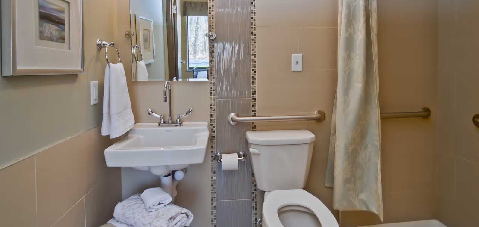 A clean and accessible bathroom featuring a white sink with a chrome faucet, a white toilet with a grab bar beside it, a towel ring with a white towel, a shower curtain with a subtle pattern, and a mirror above the sink reflecting part of the room and a window.