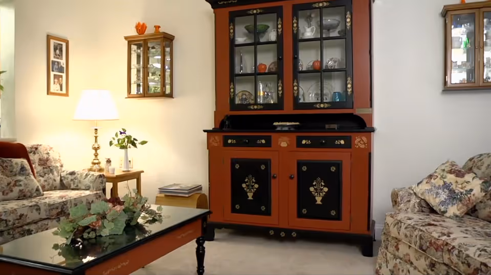 Cozy living room with floral upholstered sofas, a red and black china cabinet, glass-topped coffee table with floral arrangement, and a lit side lamp.