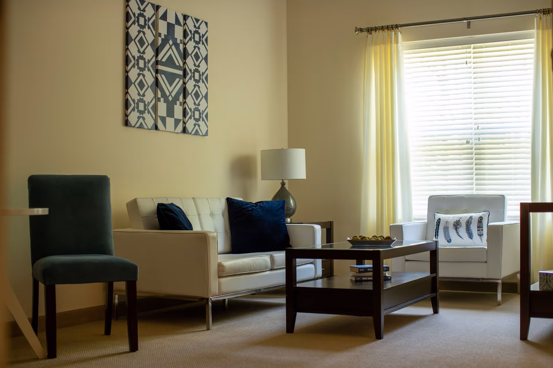 A cozy living room with a white sofa adorned with dark blue pillows, a white armchair with a decorative pillow featuring feather designs, a dark wooden coffee table with books and a decorative bowl, a table lamp on a side table, and a window with white blinds and light yellow curtains. A patterned wall art hangs above the sofa.