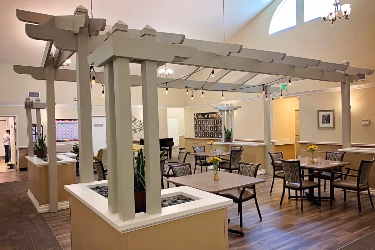 Interior of a senior living facility dining area with several tables and chairs arranged neatly. There is a decorative pergola structure in the center with string lights hanging from it. The room has wood flooring, beige walls, and high windows letting in natural light. A 'Wall of Honor' display is visible on the far wall, and a piano is placed near the back. A person is seen standing in a doorway on the left side of the image.