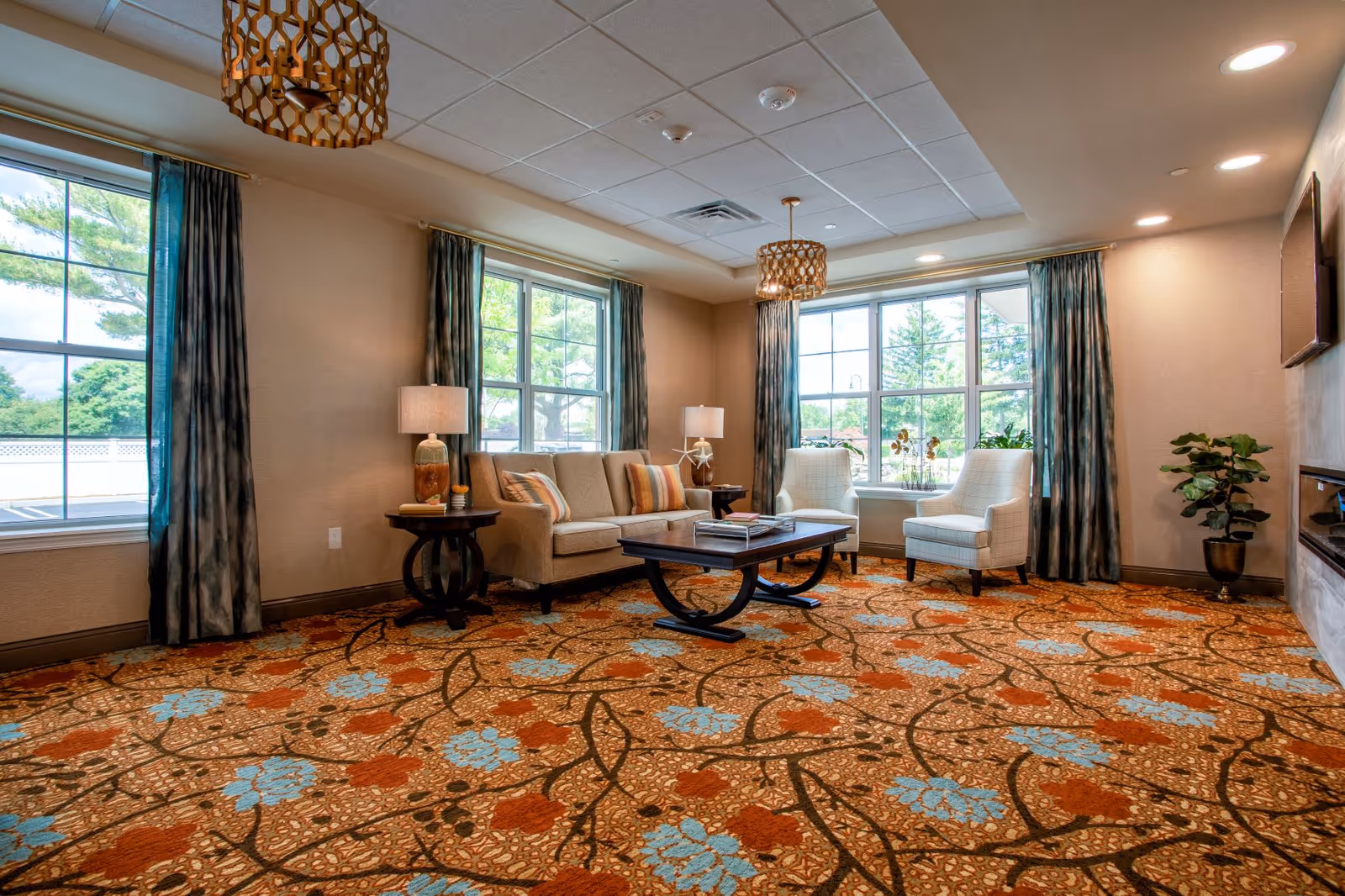 A cozy living room with a patterned carpet featuring orange and blue floral designs. The room has a beige sofa with striped pillows, two white armchairs, two side tables with lamps, large windows with blue curtains letting in natural light, and a coffee table with books on it. There is a potted plant in the corner and modern ceiling lights.