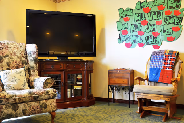 A cozy living room area with a floral upholstered armchair on the left and a wooden rocking chair with a colorful patchwork quilt draped over the back on the right. A large flat-screen TV sits on a wooden cabinet with glass doors in the center. On the wall above the rocking chair is a decorative green board with red apple shapes and various names written on it.