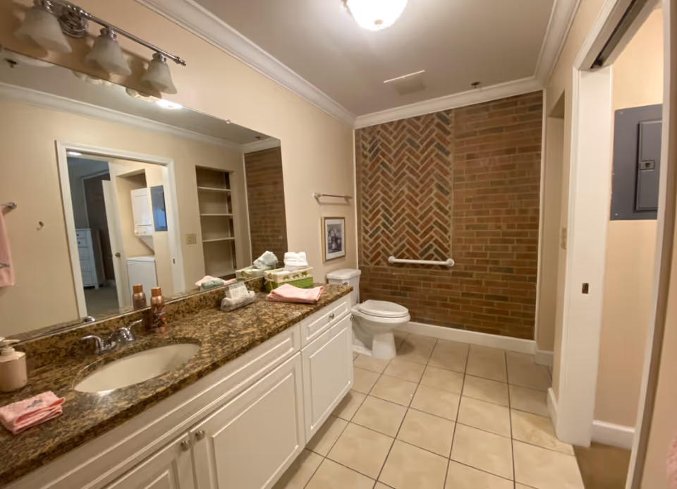 Bathroom with a long granite vanity and sink, large mirror, toilet next to a brick accent wall with a grab bar, and tiled floor.