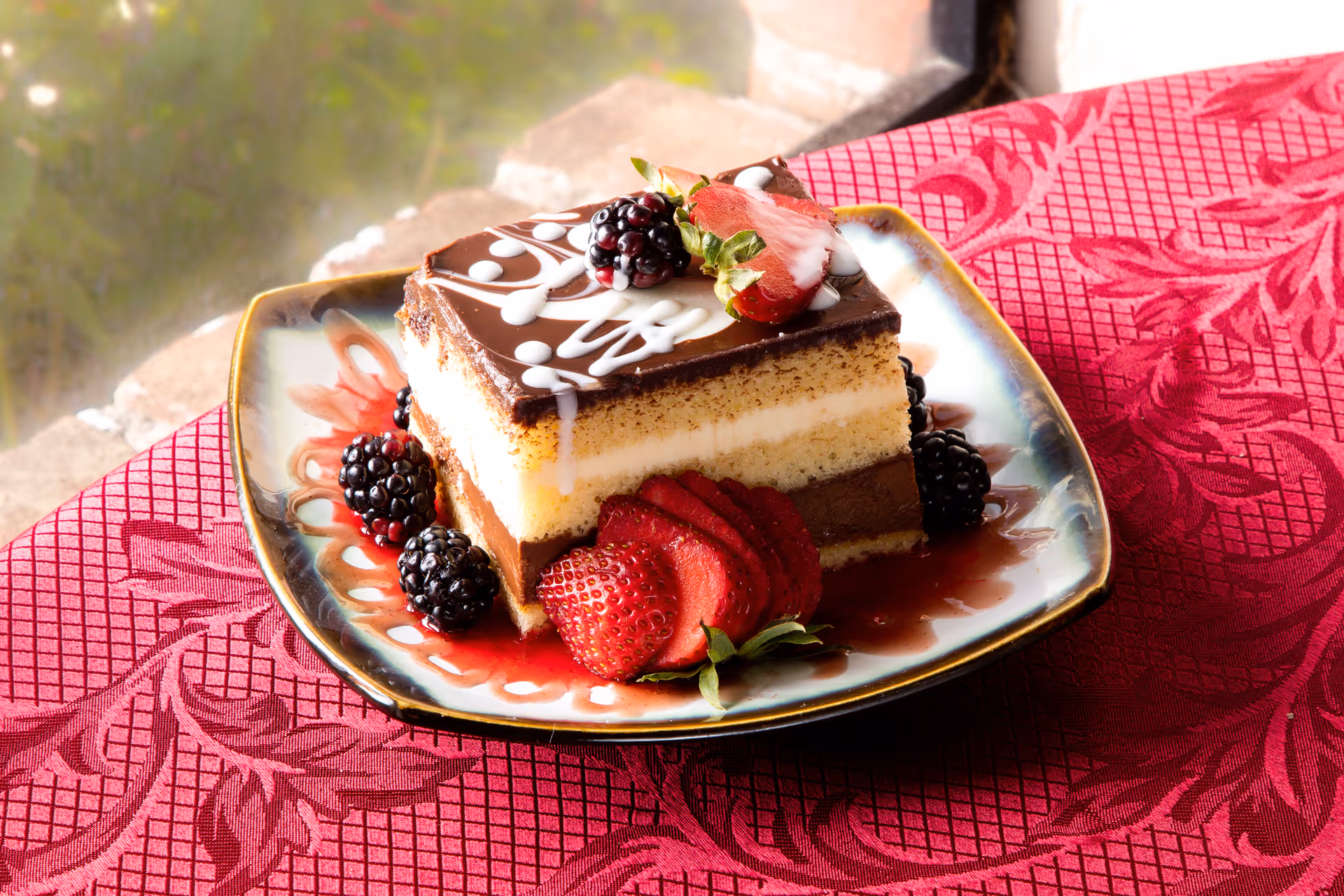 A slice of layered chocolate and vanilla cake decorated with white icing on top, served on a decorative plate with fresh strawberries and blackberries, placed on a red patterned tablecloth near a window.