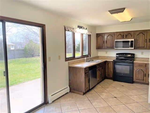 A kitchen with wooden cabinets, a black stove, microwave, dishwasher, and a double sink under a window. There is a sliding glass door leading outside to a grassy yard with a fence.
