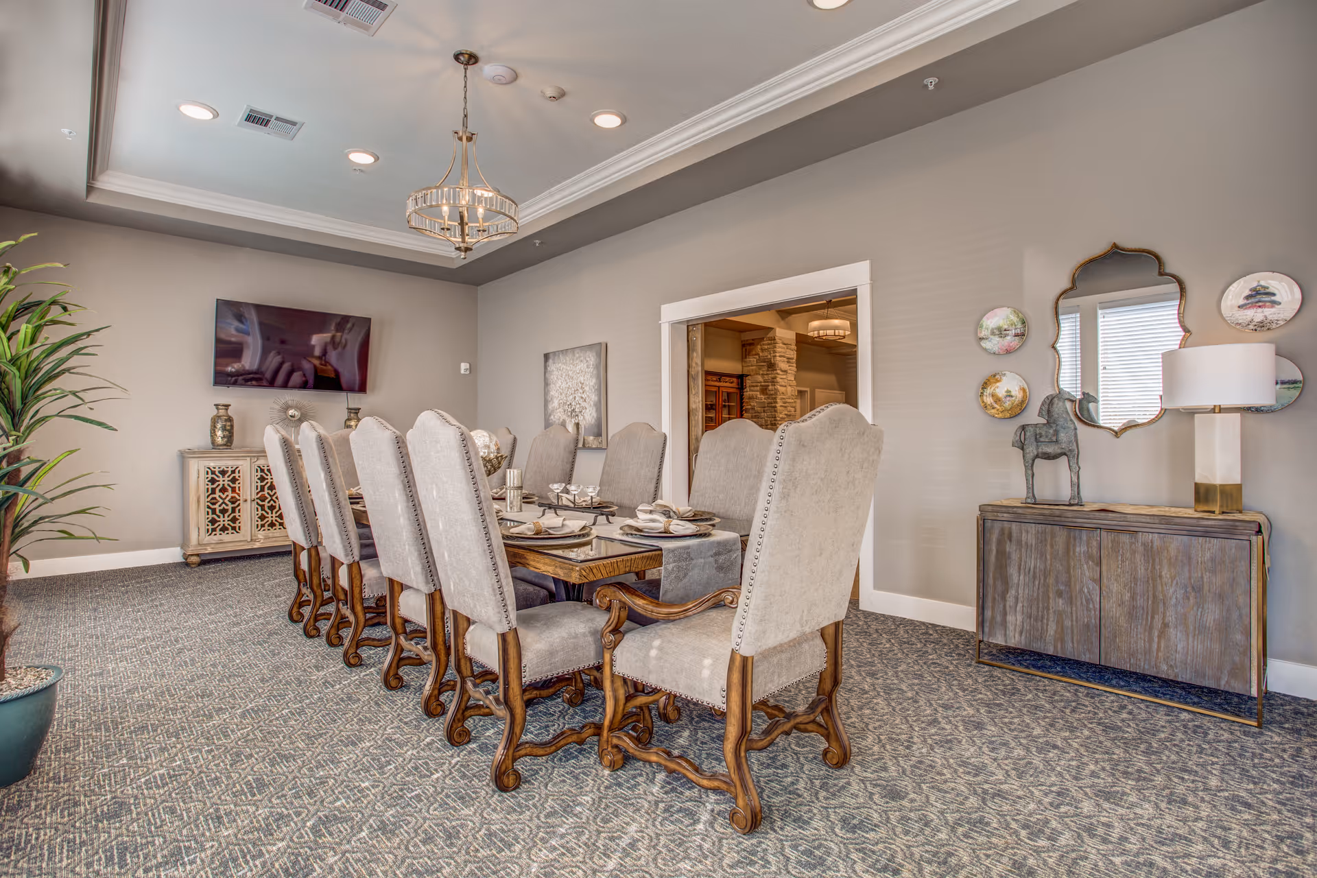A formal dining room with a long wooden table set for eight people with upholstered chairs. The room features a chandelier, a large potted plant, a wall-mounted TV, decorative plates and a mirror on the wall, and a sideboard with a lamp and horse sculpture. The carpet has a patterned design and the walls are painted light gray.