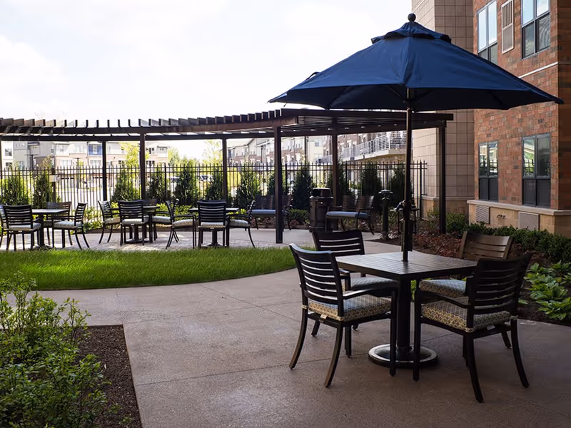 Outdoor courtyard patio with a table and chairs under a large navy umbrella, additional seating, a pergola, and landscaping beside a brick building.