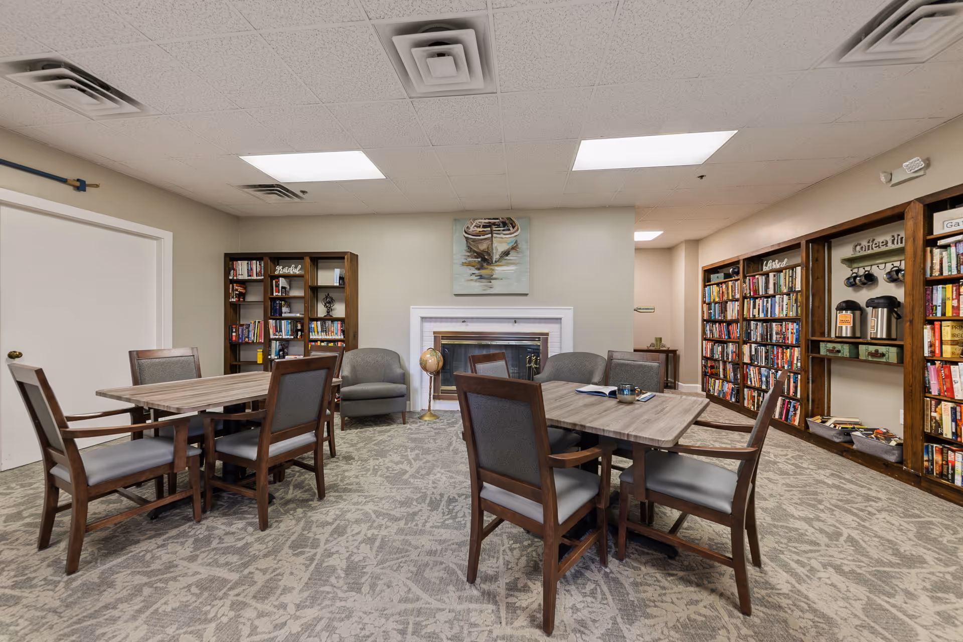 A cozy common area with two wooden tables surrounded by chairs, a fireplace with a painting of a boat above it, bookshelves filled with books, and a small coffee station with mugs and coffee pots.