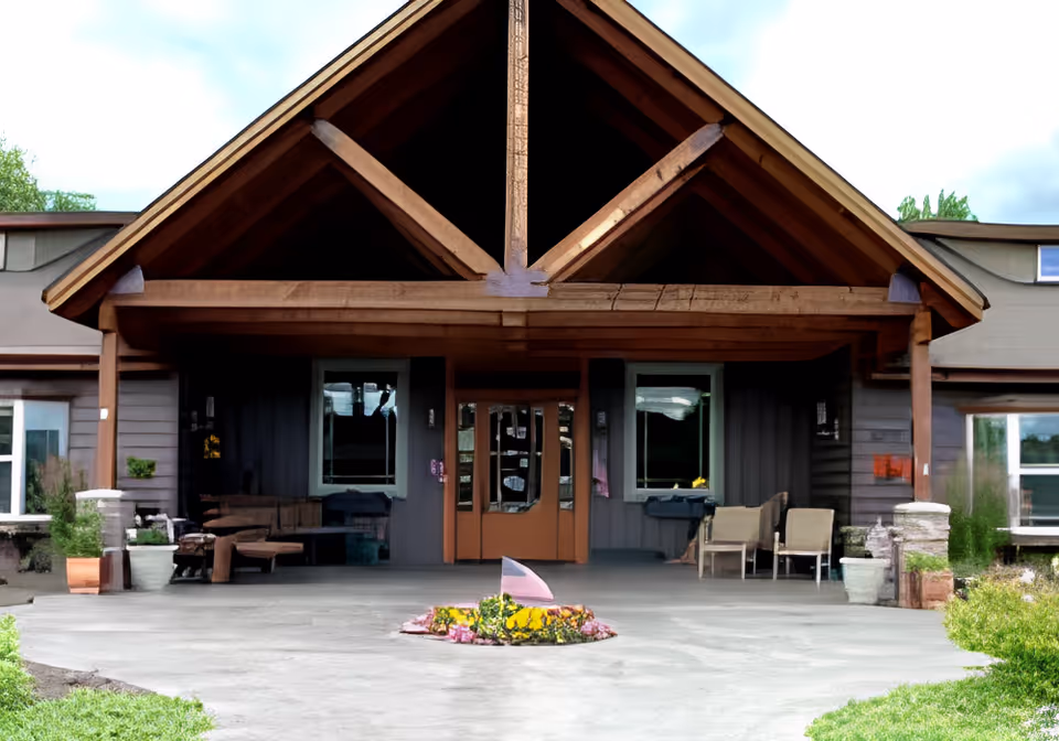 Front exterior view of Wellspring Meadows Assisted Living facility featuring a large wooden peaked entrance with glass double doors, seating areas on both sides, potted plants, and a small flower bed in the center of the concrete driveway.