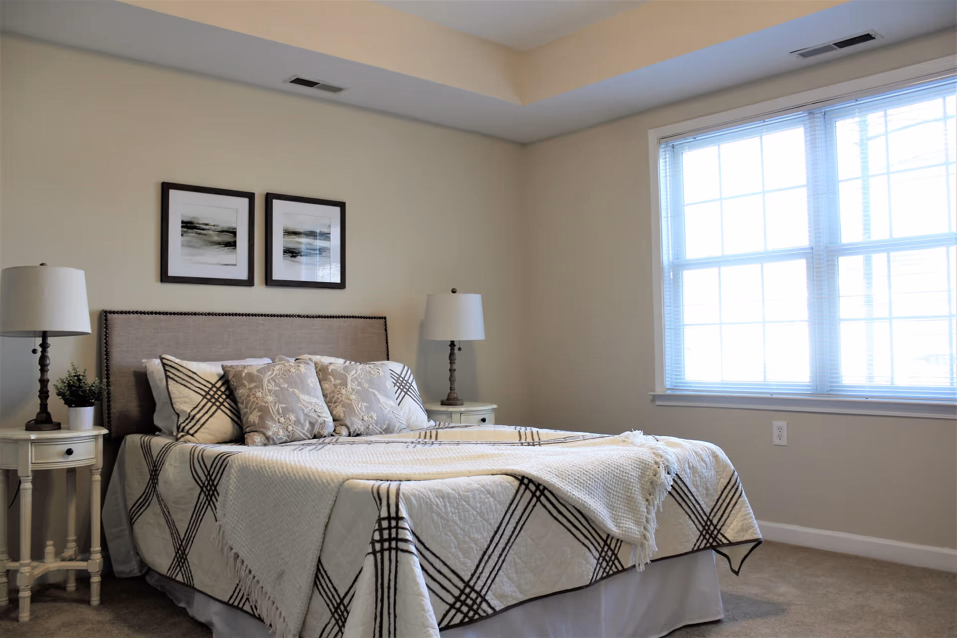 A bright bedroom with a large window on the right letting in natural light. The bed has a beige upholstered headboard and is covered with a white quilt featuring a black plaid pattern, along with several decorative pillows. On each side of the bed are white nightstands with matching lamps and a small plant on the left nightstand. Two framed abstract artworks hang on the wall above the headboard.