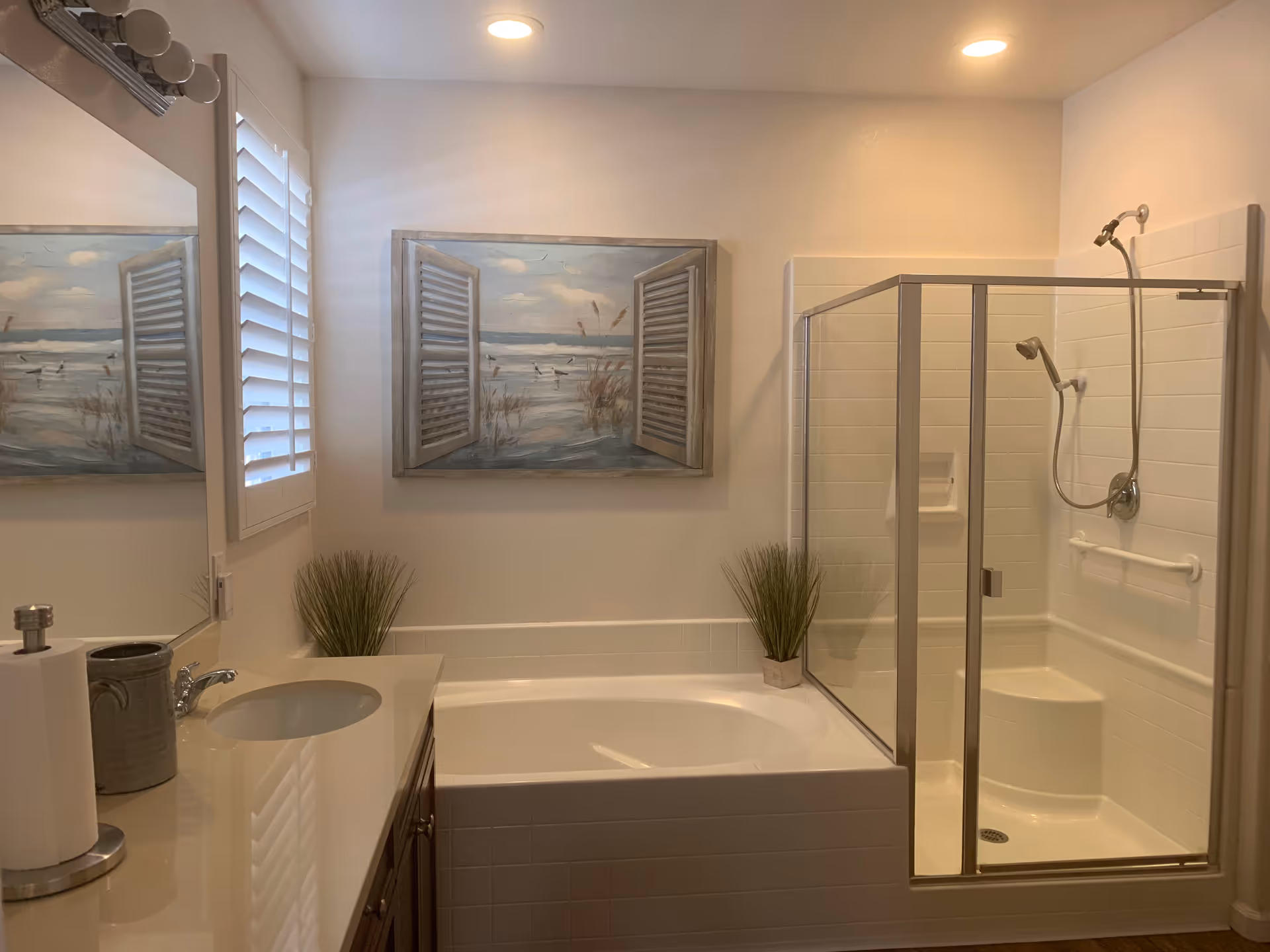 A clean and modern bathroom featuring a white bathtub with a glass-enclosed shower next to it. There is a countertop with a sink, a paper towel holder, and a decorative container. The walls are light-colored with a window covered by white shutters and a painting depicting an open window with a beach scene. Two small potted plants are placed on the bathtub ledge.