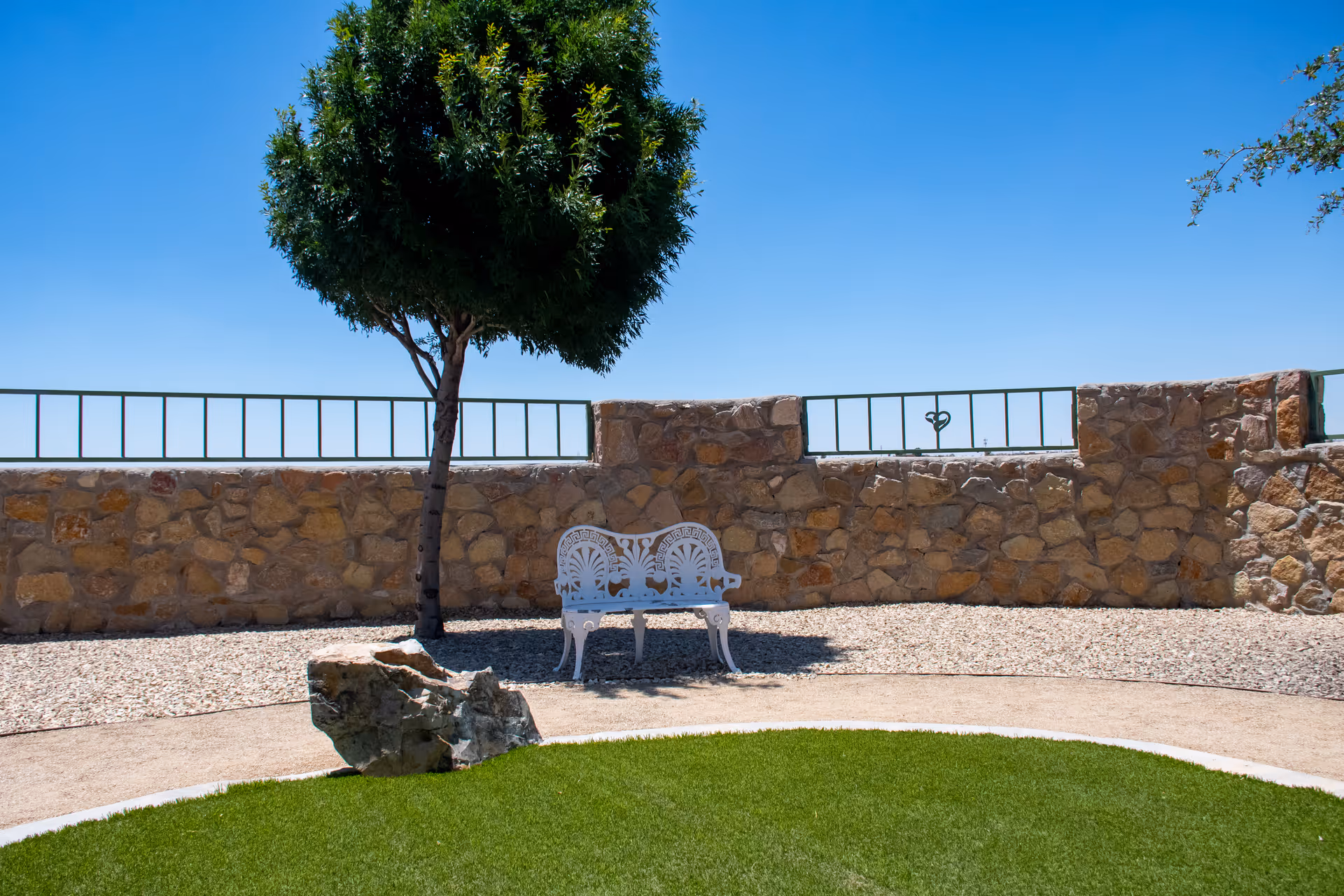 Outdoor seating area with a white decorative metal bench under a tree, surrounded by a stone wall and gravel ground, with a patch of green grass in the foreground under a clear blue sky.
