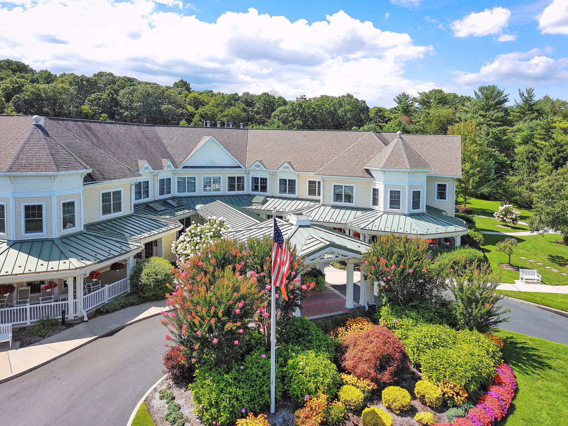 Two-story senior living building with a covered entrance, circular driveway, American flag, and colorful landscaped flower beds under a blue sky.