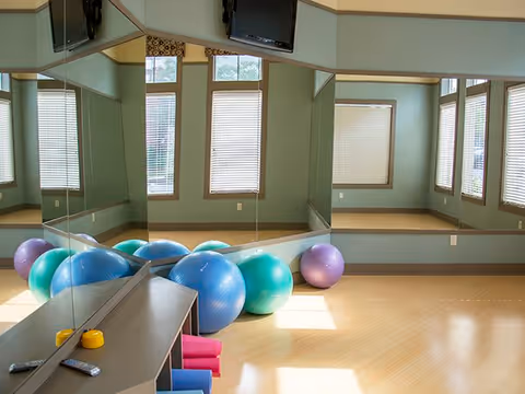 A bright exercise room with large mirrors on two walls, several colorful exercise balls on the floor, and a mounted TV screen. The room has light wooden flooring and multiple windows with blinds allowing natural light to enter.
