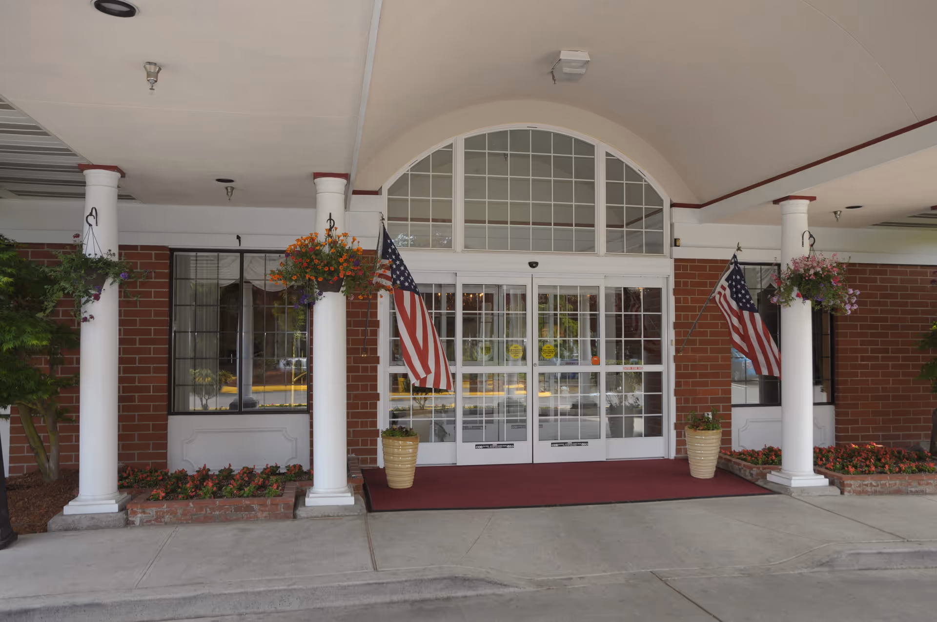 Entrance of Hallmark Manor featuring a covered driveway with white columns, hanging flower baskets, two American flags, and glass double doors with a large arched window above. There are flower beds on either side of the entrance and a red carpet leading to the doors.