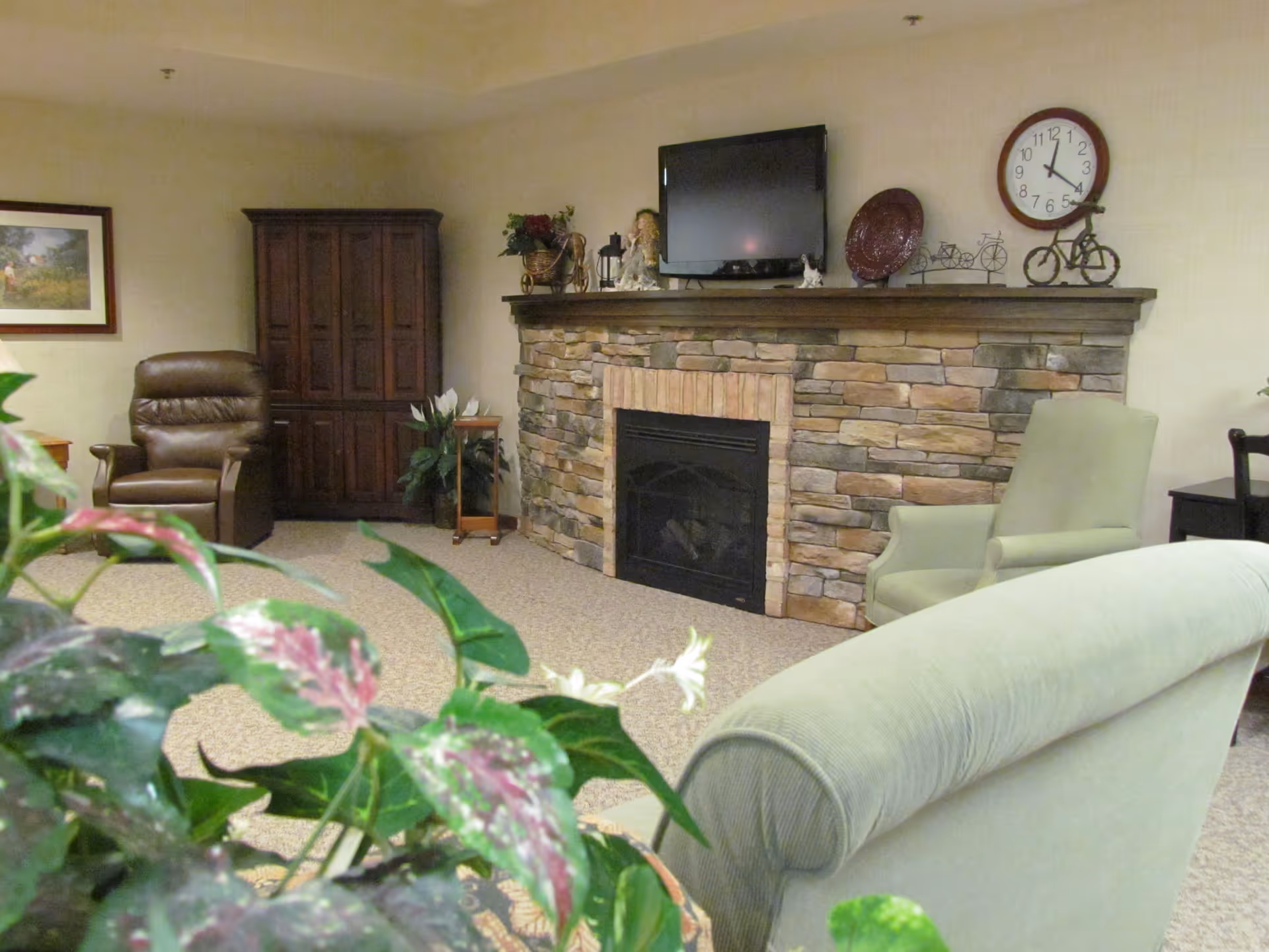 Cozy common living room featuring a stone fireplace with a mounted TV, armchairs, and potted plants.