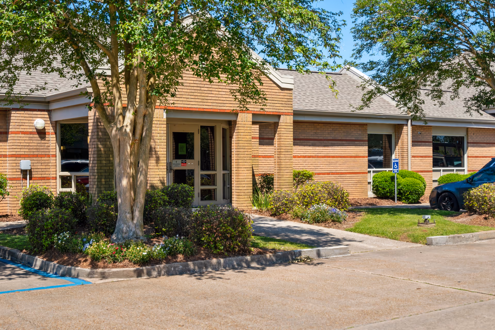 Exterior view of a single-story brick building with a main entrance door surrounded by windows, landscaped bushes, a tree, and a parking area with a handicap parking sign and a parked car.