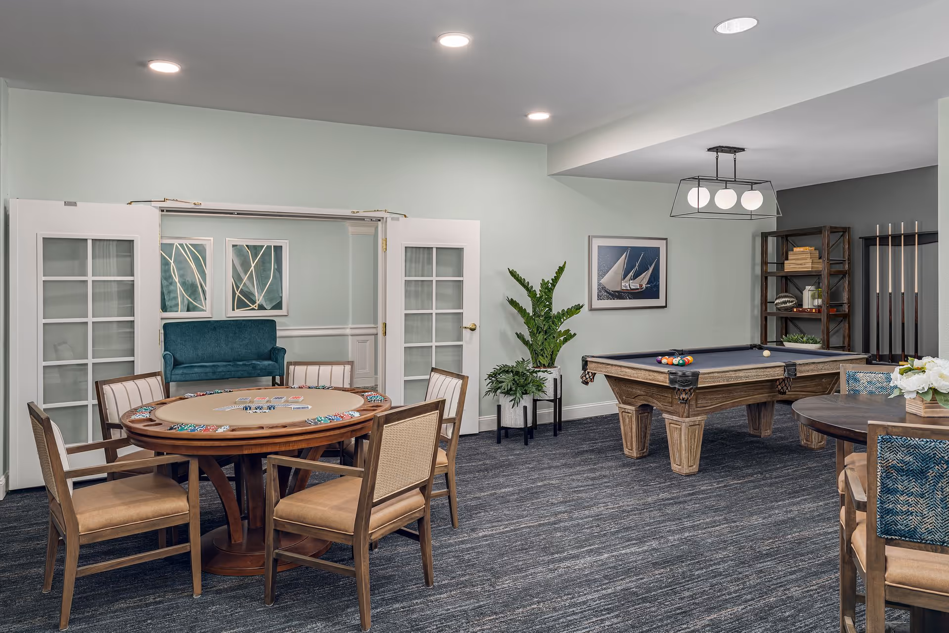 A recreational room featuring a poker table with chairs, a pool table with billiard balls, a round table with chairs, and decorative plants. The room has light-colored walls, carpeted flooring, framed artwork, and modern ceiling lights.
