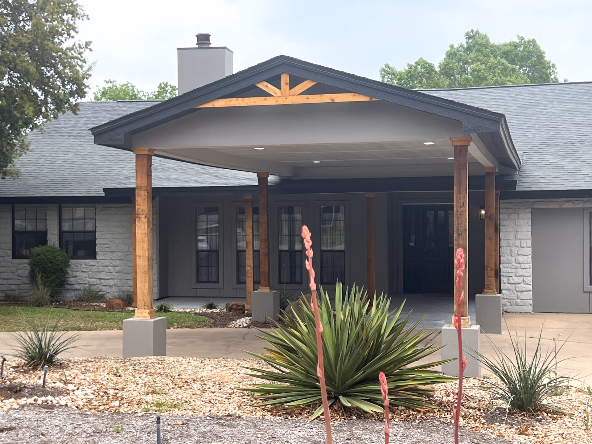 Front entrance of a single-story building with a covered portico supported by wooden posts and landscaped plants in the foreground.