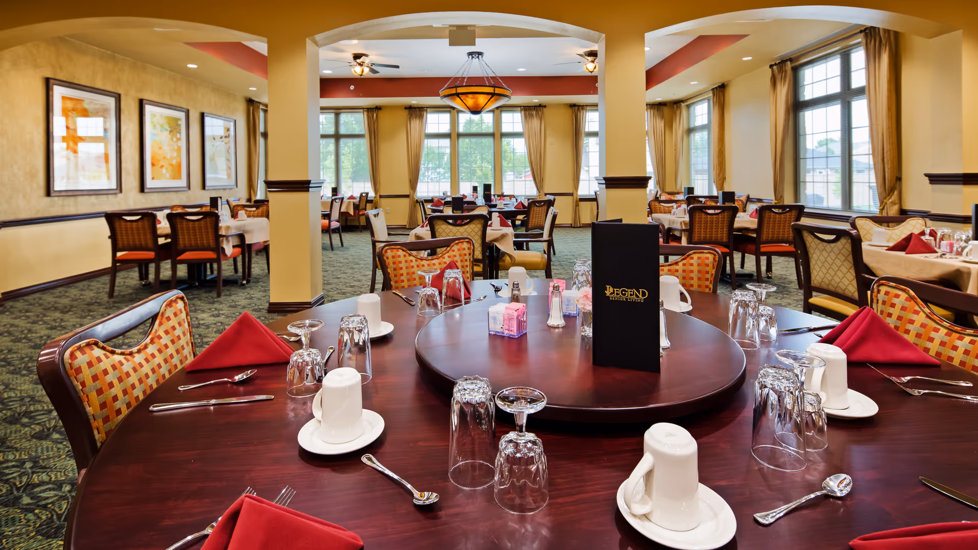 Spacious dining room with a round table in the foreground set with glasses, red napkins and a menu, and multiple tables by large windows in the background.