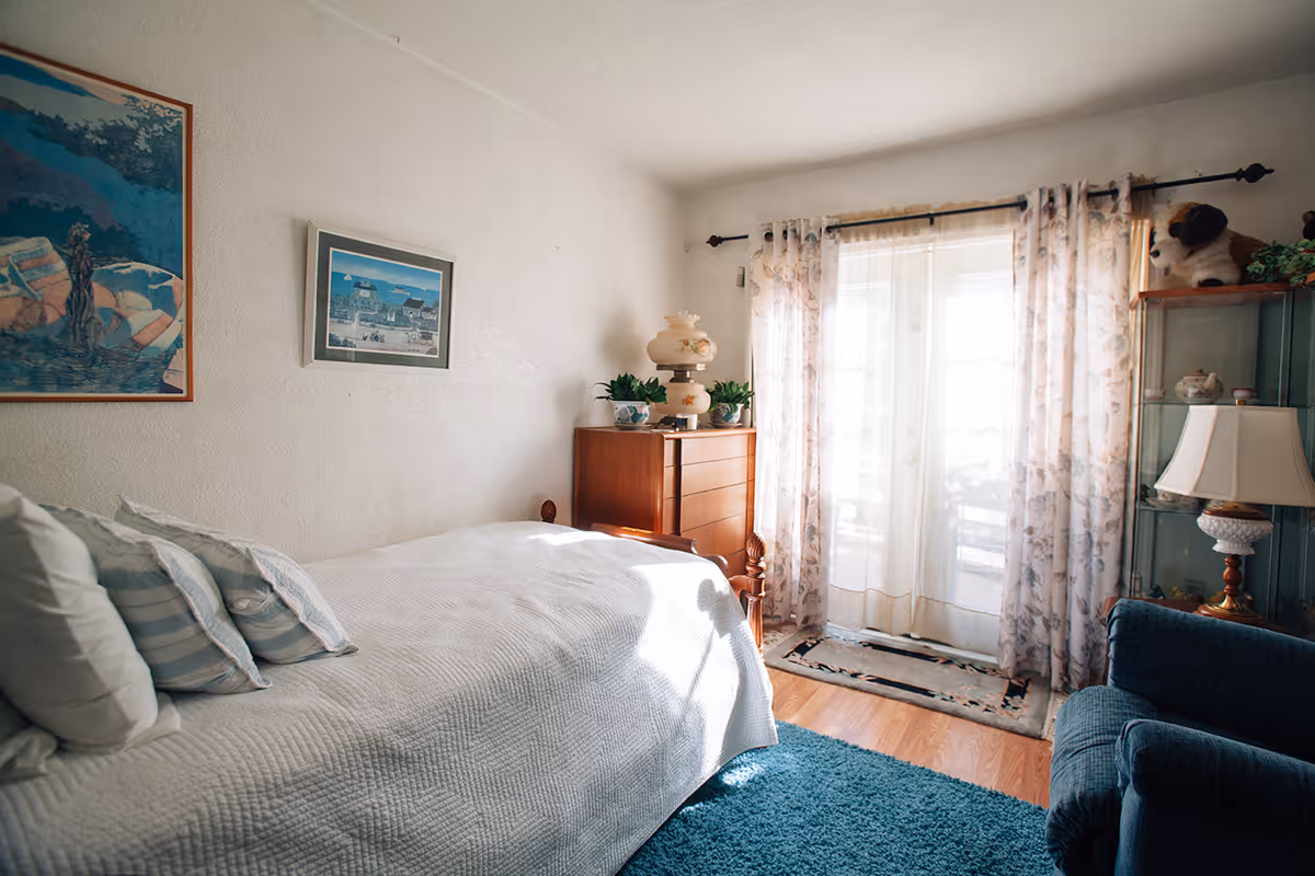 A cozy bedroom with a bed covered in a white quilt and several pillows, a wooden dresser with plants and a lamp on top, floral curtains covering glass doors, a blue armchair, a glass display cabinet with decorative items, and two framed paintings on the wall.