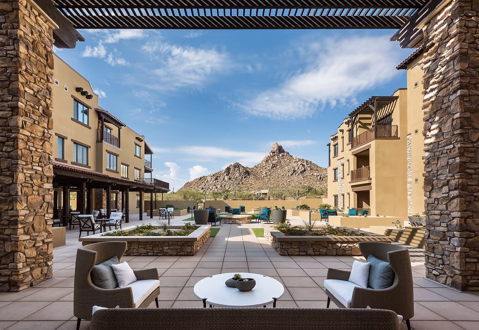 Outdoor courtyard area of ACOYA Troon senior living facility with comfortable seating, stone pillars, and a view of rocky mountains under a partly cloudy sky.