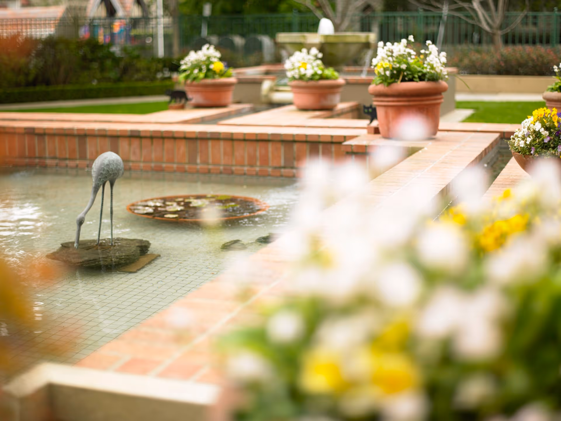 Brick-lined reflecting pool with a heron statue and potted flowers in a landscaped courtyard.