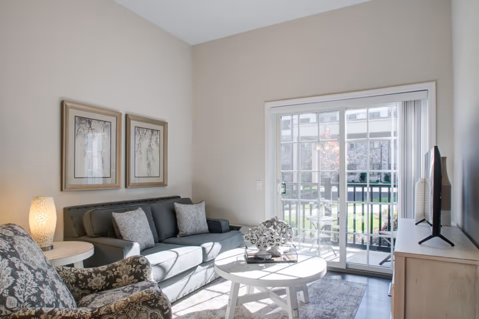 A bright living room with a gray sofa adorned with cushions, a patterned armchair, a round white coffee table with decorative items, a side table with a glowing lamp, two framed artworks on the wall, and a large sliding glass door leading to an outdoor patio area.