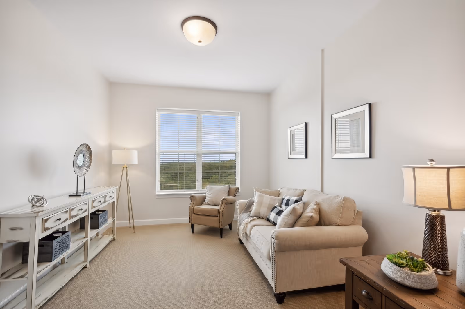 A bright living room with a beige sofa adorned with multiple cushions, a matching armchair near a window with white blinds, a white console table with decorative items, a floor lamp, two framed pictures on the wall, and a wooden side table with a lamp and a small plant.