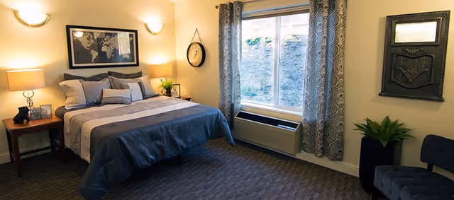 A neatly made bed with gray and beige bedding in a bedroom. There are two wooden nightstands on either side of the bed, each with a lamp. Above the bed hangs a framed world map. A window with patterned curtains lets in natural light, and below it is an air conditioning unit. A round wall clock hangs next to the window, and a potted plant is placed near a dark upholstered chair in the corner.