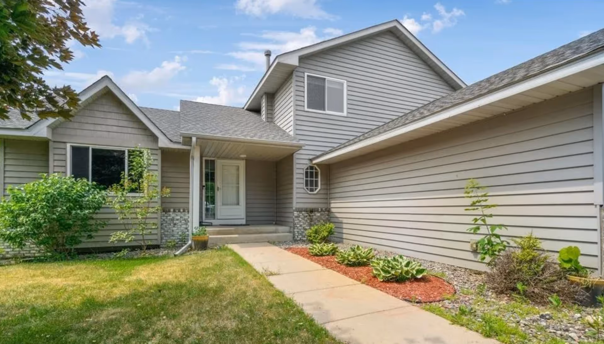 Front entrance of a gray-sided residential building with a concrete walkway, small garden beds and a covered porch.