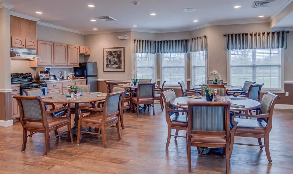Dining area with several round tables and wooden chairs, a small kitchenette to the left, and large windows with valances.